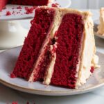 A homemade gluten free red velvet cake slice with deep red, airy layers and creamy, off-white frosting. The frosting is swirled and slightly uneven, giving it a rustic look. Crumbs are scattered on the plate and cake stand, with another slice slightly blurred in the background. The soft lighting enhances the cake’s rich color and moist texture.