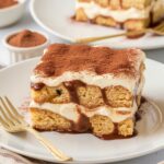 A homemade slice of gluten free tiramisu served on a white plate with a gold fork beside it. The dessert consists of two layers of coffee-soaked gluten-free ladyfingers, enveloped in a rich mascarpone cream filling. The mascarpone mixture is slightly melting over the edges, creating an irresistible, creamy texture.