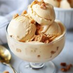 A close-up of creamy homemade dulce de leche ice cream in a glass dish, topped with golden crumbles. The smooth scoops slightly drip down the sides, and a golden spoon is visible in the background.