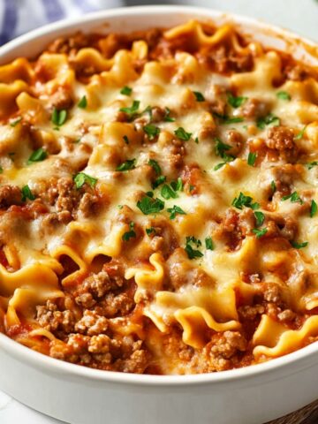 A homemade Hamburger Helper lasagna in a round white dish, topped with melted mozzarella, browned ground beef, and a sprinkle of fresh parsley. Curly lasagna noodles peek through a rich tomato meat sauce, with a blue-striped towel in the background on a white countertop.