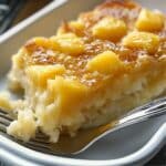 A moist, homemade pineapple dump cake slice on a white plate, topped with caramelized pineapple chunks and a glossy, golden-brown glaze, with a fork cutting into its fluffy, dense texture.