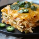 A slice of homemade chicken enchilada casserole on a black plate, topped with melted cheddar cheese, cilantro, and jalapeño slices. Layers of shredded chicken, tortillas, and veggies are visible, with extra jalapeños on the plate.