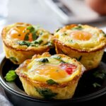 Three golden-brown homemade Egg Bites Muffin Tins with whole egg yolks, herbs, and veggies, served in a black dish on a white napkin, with eggs and a glass of water in the blurred background.