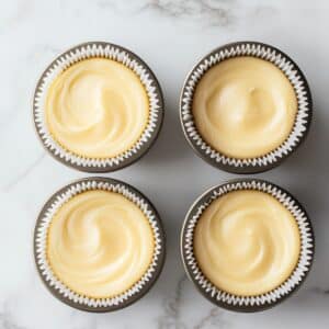 Four unbaked lemon cupcakes in a dark non-stick muffin tin, each filled with smooth, swirled yellow batter in white paper liners on a marble countertop.