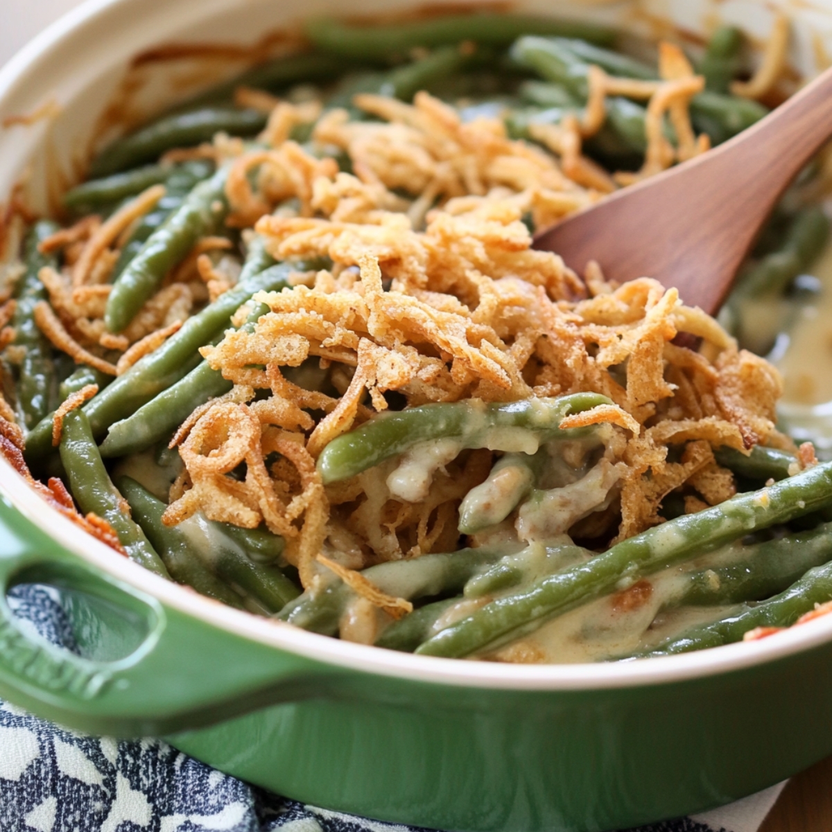 Homemade green bean casserole in a green ceramic dish, topped with golden crispy onions and creamy mushroom sauce, with a wooden spoon ready to serve.