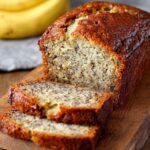 Homemade Banana Bread Recipe loaf with a golden-brown crust and two moist slices cut on a wooden board, with ripe bananas in the background.