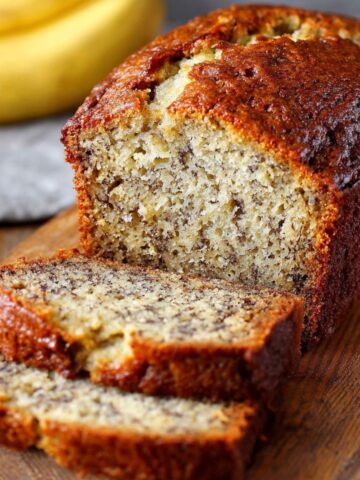 Homemade Banana Bread Recipe loaf with a golden-brown crust and two moist slices cut on a wooden board, with ripe bananas in the background.