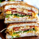 Homemade triple-decker club sandwich with toasted bread, turkey, ham, bacon, lettuce, tomato, scrambled eggs, cheddar cheese, and mayo, sliced and stacked on a wooden board with toothpicks.