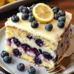 A homemade slice of lemon blueberry cake with white glaze, fresh blueberries, and a lemon slice on top, served on a white plate with a fork.