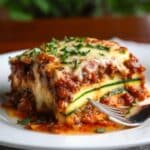 A slice of homemade zucchini lasagna with melted cheese, meat sauce, and zucchini layers on a white plate, garnished with parsley and served with a fork.