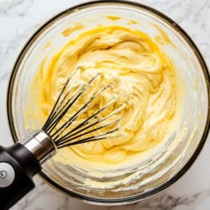 Lemon cupcake batter being mixed in a clear glass bowl with a handheld whisk, showing a smooth, creamy texture and swirls on a marble countertop.