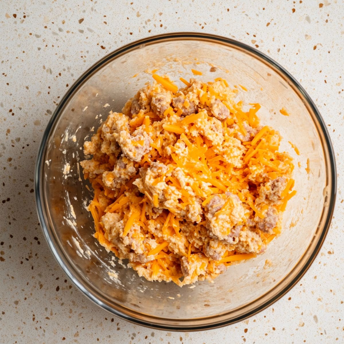 Unmixed sausage ball dough with shredded cheddar, sausage, and biscuit mix in a glass bowl on a speckled countertop.