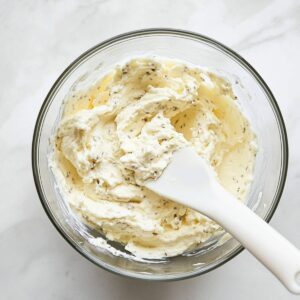 Creamy ricotta cheese and herb mixture in a glass bowl with a white spatula on a marble surface.