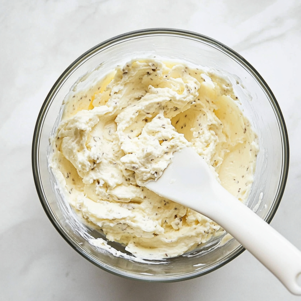 Creamy ricotta cheese and herb mixture in a glass bowl with a white spatula on a marble surface.