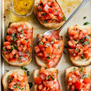 Toasted bread slices topped with fresh tomato, basil, and garlic mixture, with a spoon mid-assembly on a baking sheet.