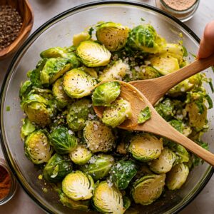 Halved Brussels sprouts in a glass bowl being tossed with wooden spoons, seasoned with coarse salt, cracked black pepper, and spices, coated in olive oil in a bright, home kitchen setting.