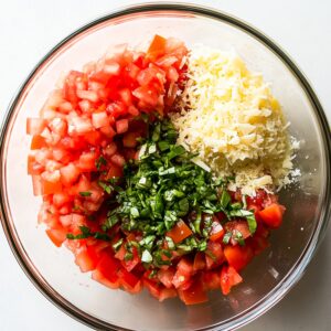 Glass bowl with diced tomatoes, chopped basil, and shredded Parmesan, ready to mix for homemade bruschetta.