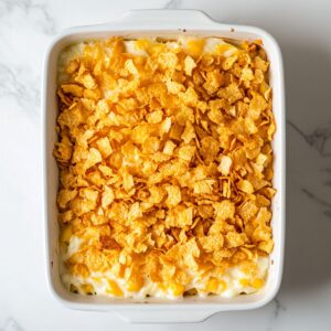 Unbaked hashbrown casserole in a white baking dish, topped with a layer of crushed golden cornflakes, sitting on a marble countertop.