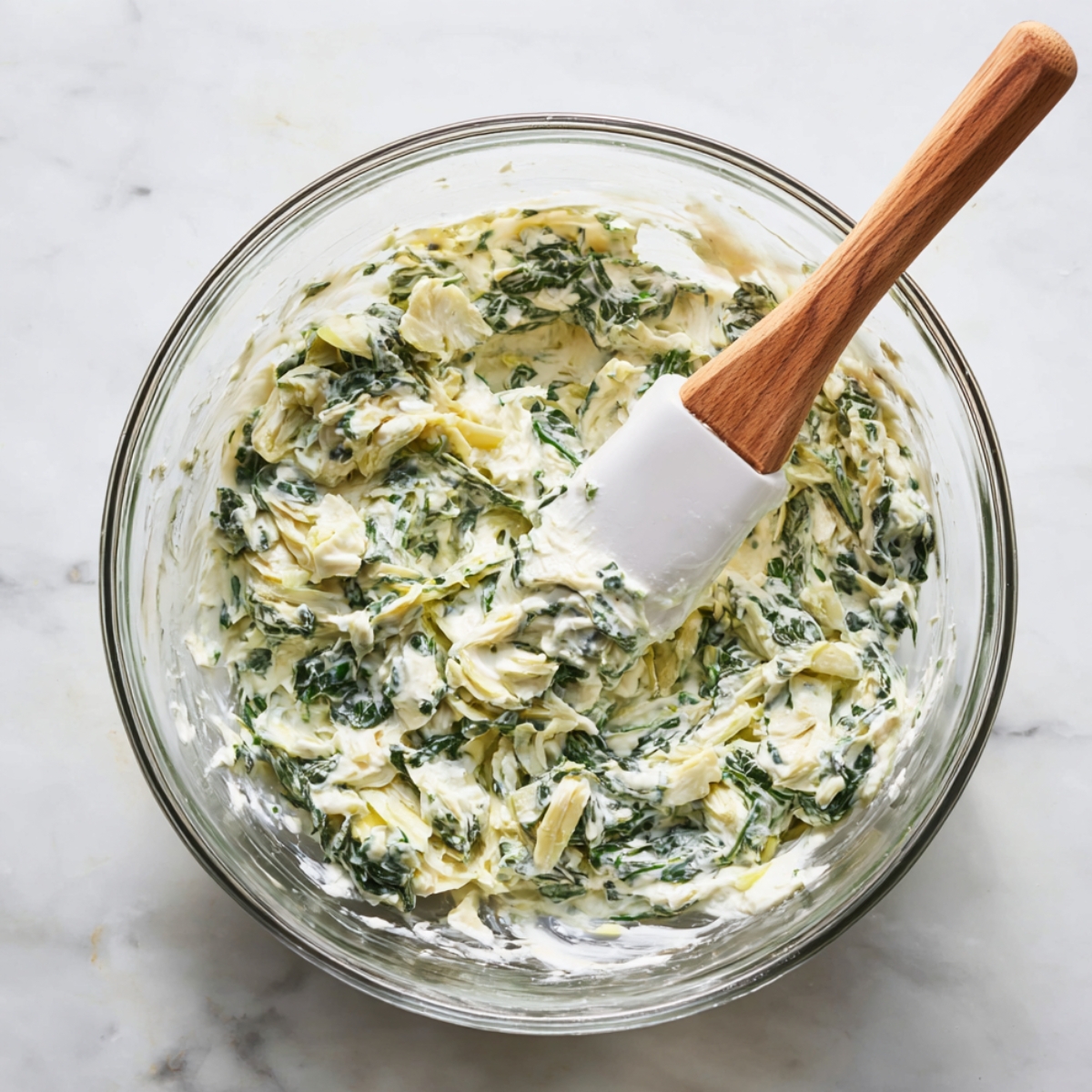 Glass bowl of creamy spinach artichoke dip with visible spinach and artichoke pieces, mixed with a white spatula on a marble counter.