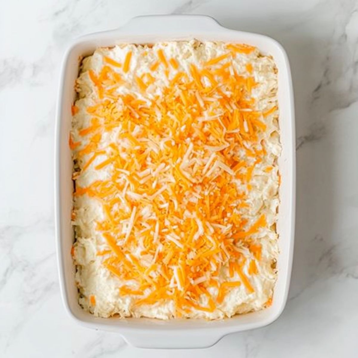 Unbaked hashbrown casserole topped with shredded cheese in a white dish on a marble countertop.