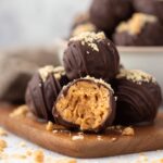 Bite-sized, homemade peanut butter balls coated in dark chocolate and drizzled with peanut butter, with one ball showing a creamy, textured peanut butter center and peanut crumbles scattered on a marble surface.