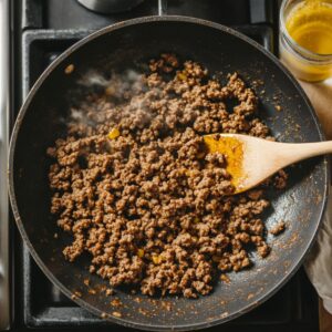 Ground beef browning in a non-stick skillet on a stovetop, stirred with a wooden spoon, steam rising as it's seasoned for taco pizza.