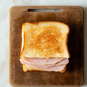 A toasted Monte Cristo sandwich with sliced ham, placed on a wooden cutting board, showing a golden, crisp top slice of bread with ham slightly sticking out.
