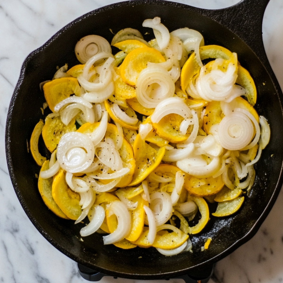 Sautéed yellow squash and sliced onions cooking in a cast iron skillet on a marble countertop, seasoned with salt and pepper.