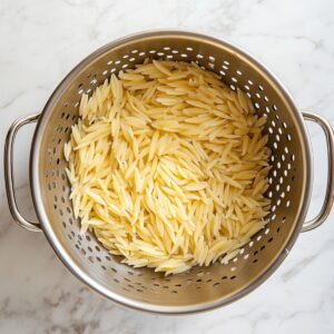 Cooked orzo pasta draining in a metal colander over a white marble countertop, freshly rinsed and ready for salad prep.