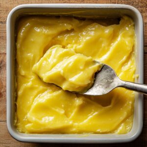 Smooth lemon pie filling being spread in a square white baking dish with a metal spoon, creating an even layer for homemade lemon cream cheese dump cake.