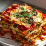 Close-up of a tall slice of homemade vegetable lasagna on a white plate, showing layers of pasta, roasted vegetables, red sauce, melted cheese, and fresh parsley on top.