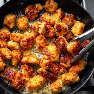 Golden-brown bite-sized chicken pieces frying in a black skillet, bubbling in oil, with metal tongs lifting one crispy piece.