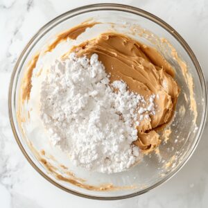 Glass mixing bowl with creamy peanut butter and powdered sugar, partially combined, on a white marble countertop—midway through making homemade peanut butter ball dough.