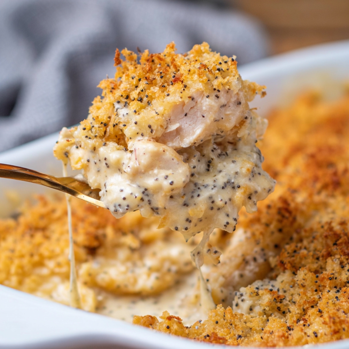 A creamy, cheesy scoop of homemade poppy seed chicken casserole with crispy golden topping being lifted from a white baking dish, showing tender chicken and melted cheese.