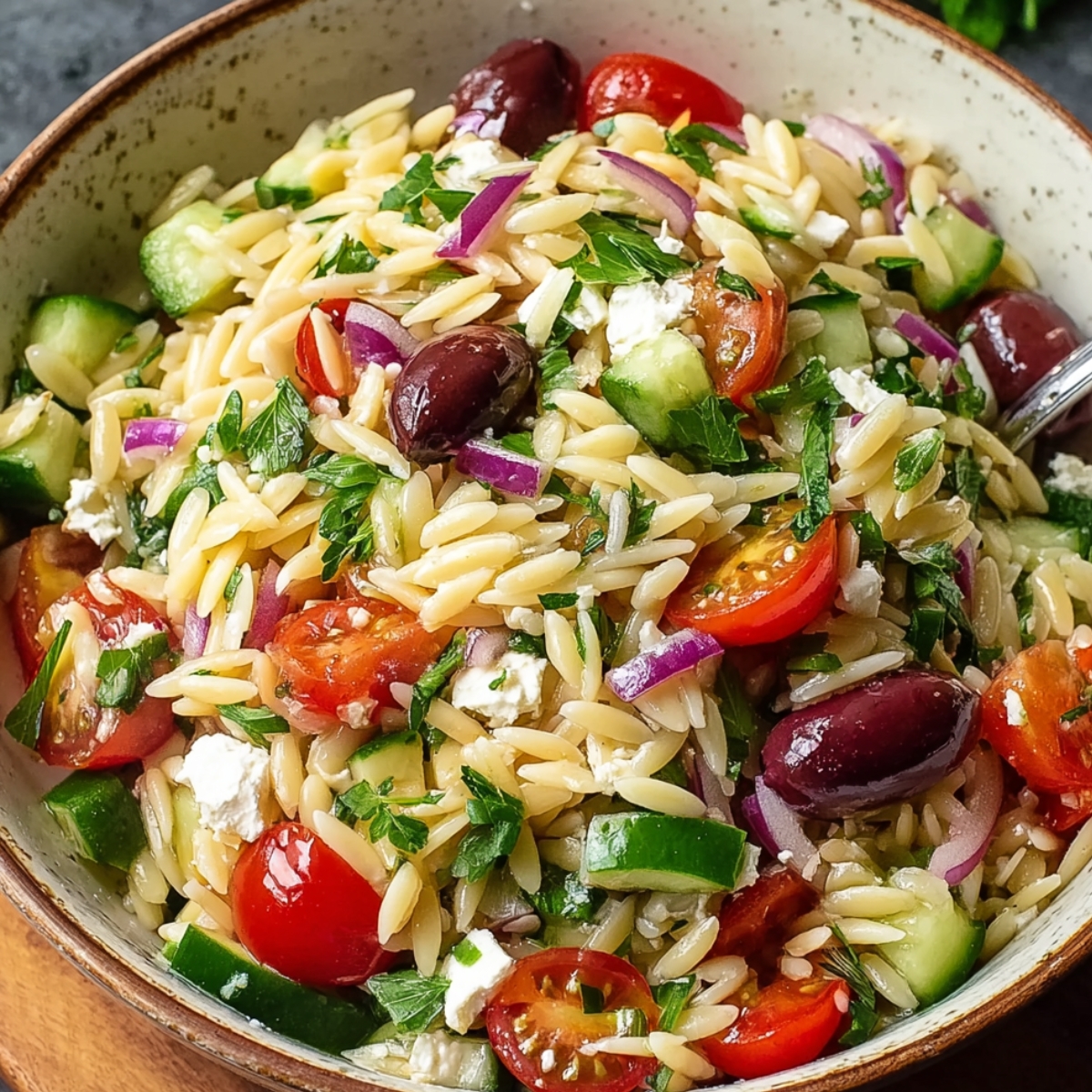 Homemade Greek orzo salad with cherry tomatoes, cucumber, red onion, Kalamata olives, feta cheese, and parsley in a rustic bowl, lightly dressed with olive oil.