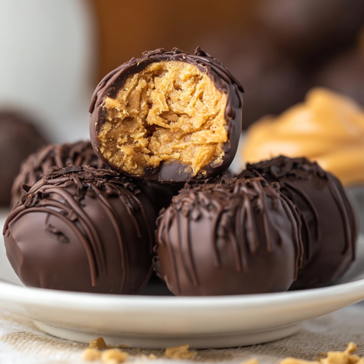 Homemade chocolate-covered peanut butter balls on a white plate, with one bitten to show the creamy, textured peanut butter filling. Drizzled chocolate and peanut crumbs add a rustic, real-kitchen touch.