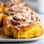 Homemade pumpkin cinnamon rolls topped with cream cheese glaze, showing golden-orange dough swirls and gooey spiced filling on a white plate.