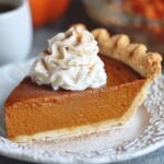 A slice of homemade pumpkin pie with a golden, flaky crust and creamy orange filling, topped with whipped cream and cinnamon, on a white plate with coffee and pumpkins in the background.