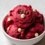 Homemade red velvet cookie dough scoops in a white bowl, studded with white and dark chocolate chips, on a light gray surface.