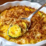 Homemade squash casserole with a golden breadcrumb topping, served in a white ceramic dish. A spoon lifts out creamy, cheesy squash slices.