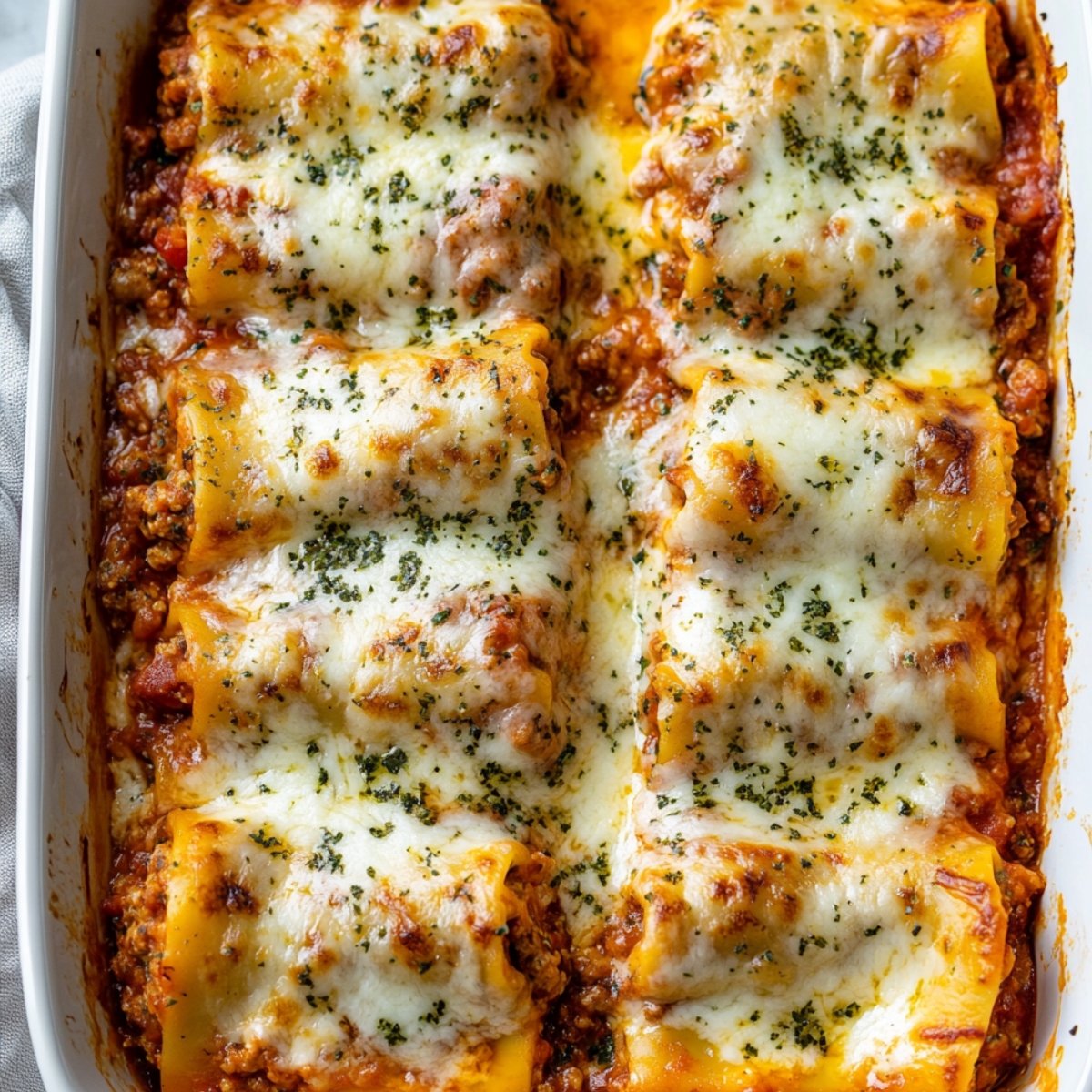 Baked lasagna roll ups in a white dish, topped with melted golden cheese and sprinkled with parsley over rich meat sauce.
