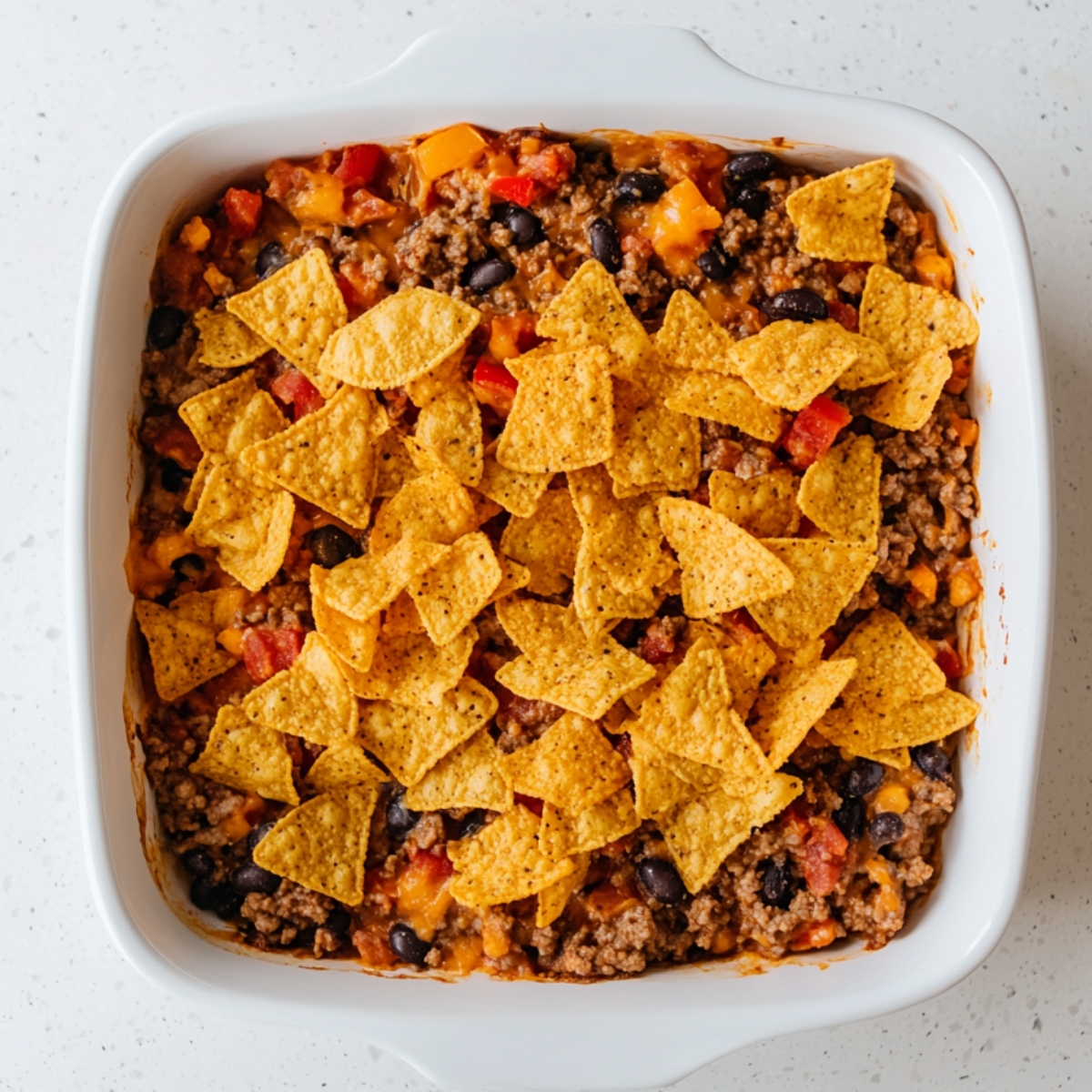 Partially assembled Dorito casserole in a white baking dish with a visible layer of seasoned ground beef, black beans, melted cheese, diced tomatoes, and a topping of whole nacho cheese tortilla chips.
