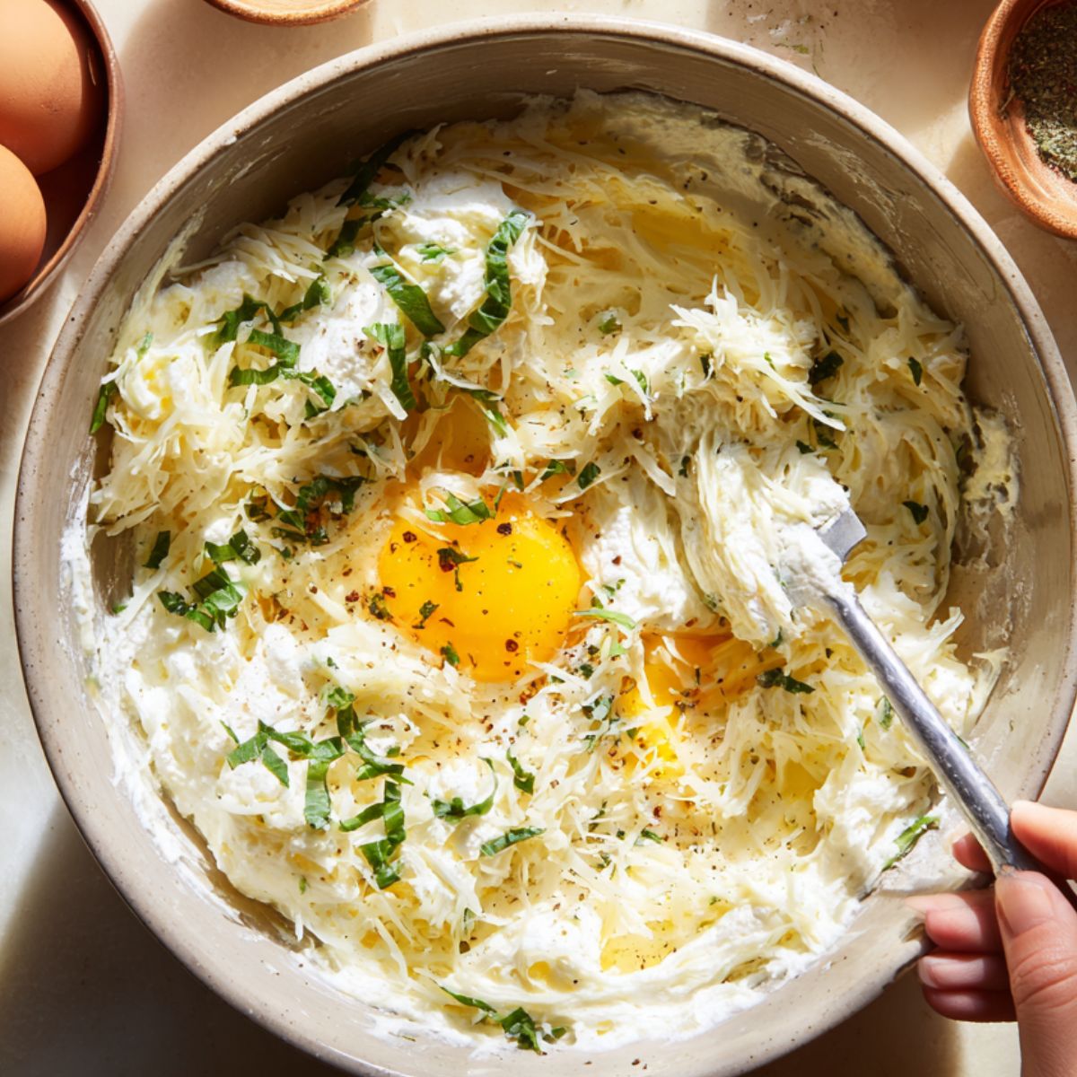 A mixing bowl with ricotta, shredded cheese, fresh basil, eggs, and black pepper being stirred by hand, making a creamy lasagna cheese mixture.