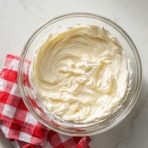 A glass bowl filled with smooth, fluffy homemade cheesecake filling, resting on a red and white checkered kitchen towel on a white marble countertop.