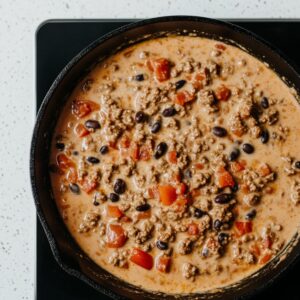 Creamy Dorito casserole filling in a skillet with ground beef, diced tomatoes, black beans, and melted cheese sauce simmering on a stovetop, ready for layering.