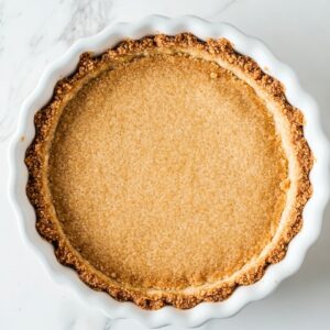 Top-down view of a homemade graham cracker pie crust in a white fluted dish, golden brown with a fine, crumbly texture, on a marble surface.
