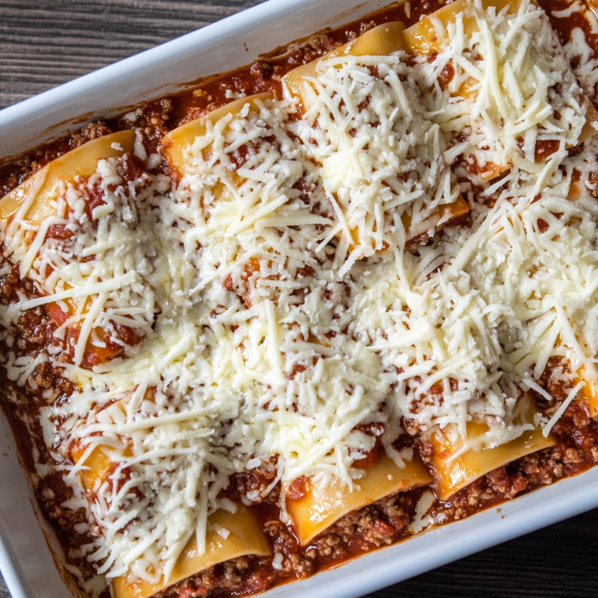Lasagna roll ups in a white baking dish, topped with shredded mozzarella and nestled in meat marinara sauce, ready to bake.