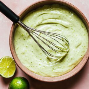 Top-down view of a bowl filled with creamy Key Lime Pie filling, pale green with flecks of lime zest. A metal whisk rests in the mixture, and fresh limes sit nearby on a light pink surface.