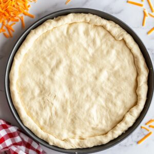 Raw pizza dough pressed into a round baking pan with raised edges, ready for taco pizza, surrounded by shredded cheddar and a red gingham cloth on a marble countertop.