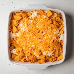 Unbaked Dorito casserole in a white square baking dish, topped with crushed nacho cheese chips and shredded cheddar over a creamy white cheese layer, ready for the oven.